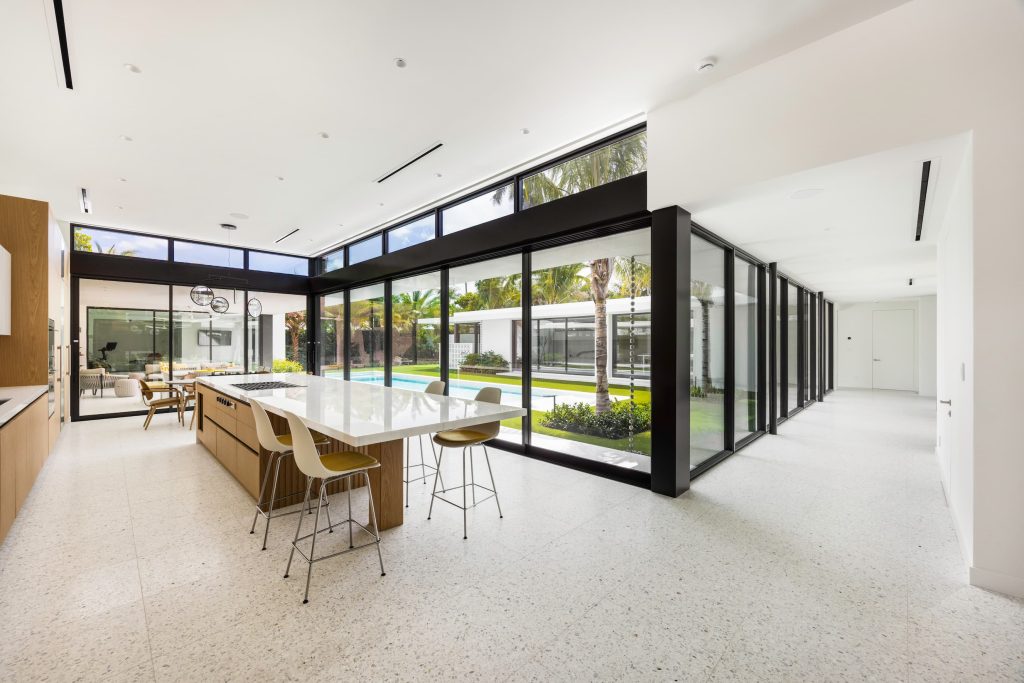 Contemporary open-plan kitchen featuring floor-to-ceiling glass walls, a central island with seating, wood cabinetry, and seamless indoor-outdoor connection to a landscaped courtyard and pool