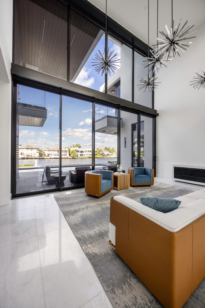 Luxury double-height living room featuring floor-to-ceiling glass walls, modern seating, sculptural pendant lighting, and expansive waterfront views through sliding glass doors