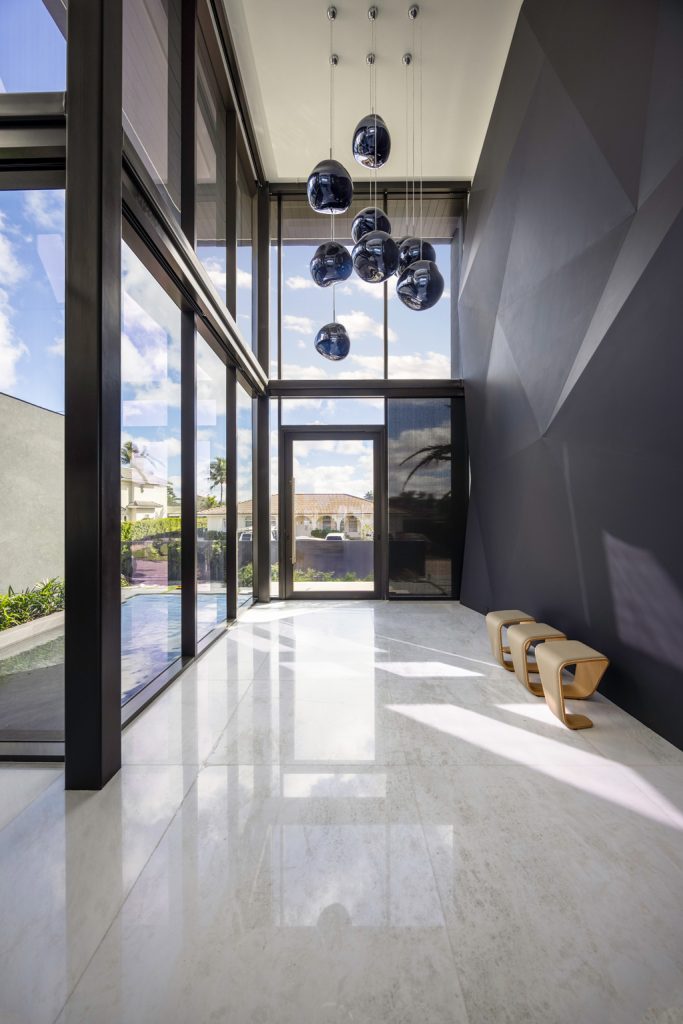 Modern double-height entryway featuring floor-to-ceiling glass walls, a sculptural pendant light installation, polished stone flooring, and abundant natural light