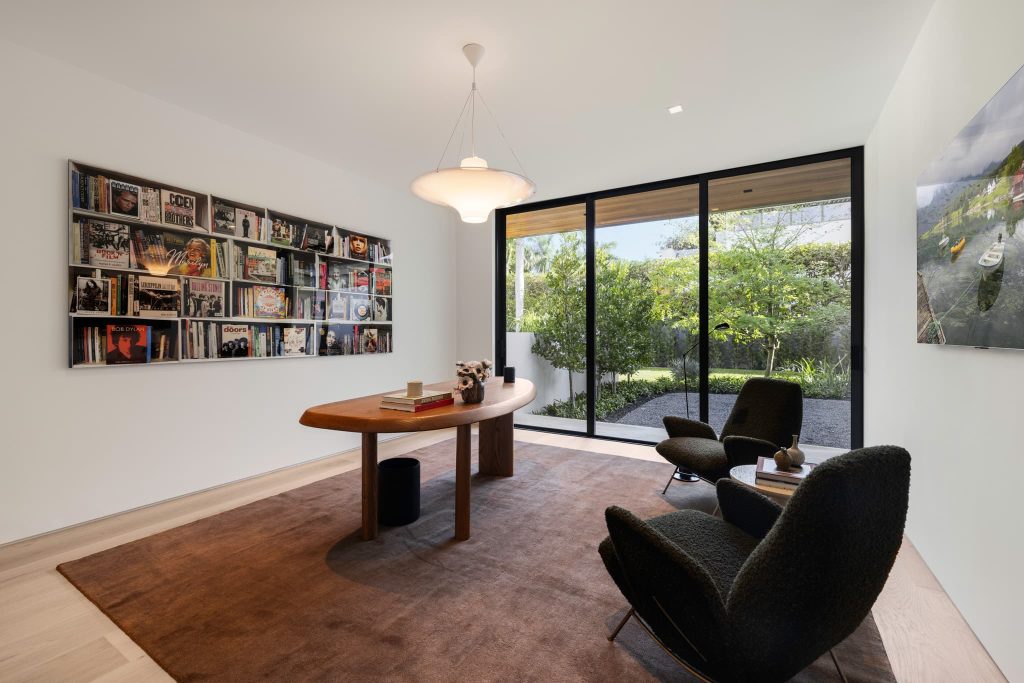 Modern home office featuring sliding glass doors, a wooden desk, lounge seating, wall-mounted books and vinyl records, and a peaceful view of a landscaped garden