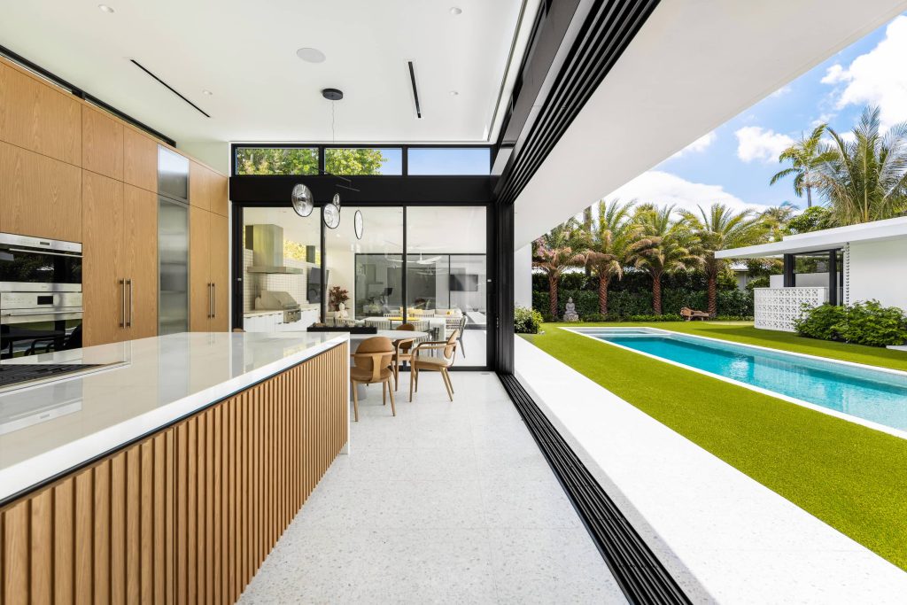 Modern open-concept kitchen featuring retractable glass walls, wood cabinetry, minimalist design, and seamless access to an outdoor pool and garden
