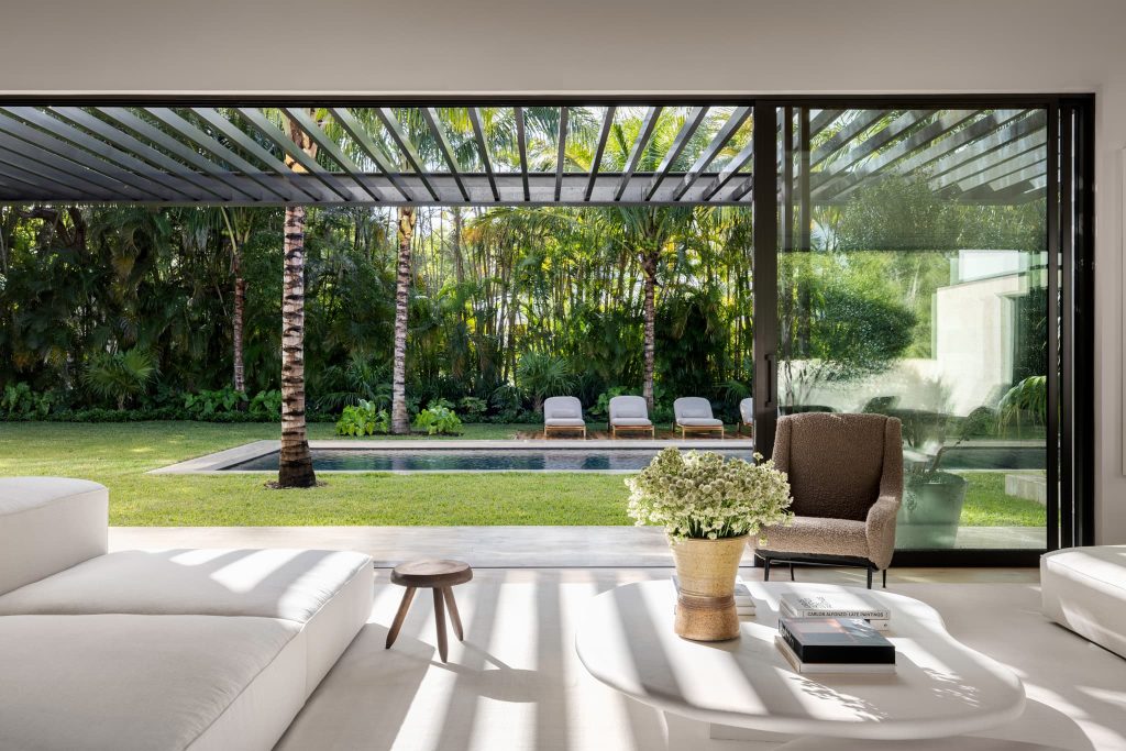 Modern living room with floor-to-ceiling sliding glass doors, minimalist furniture, and a view of a tropical garden with a pool and palm trees