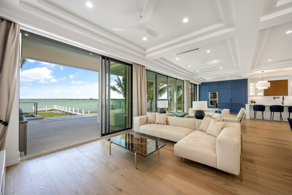 Elegant waterfront living room with large sliding glass doors, modern furniture, wood flooring, and an open view to a terrace overlooking the water